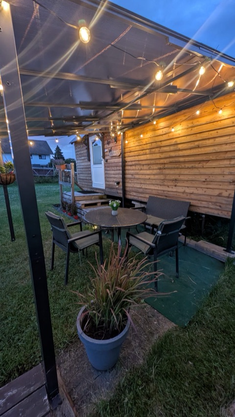 Überdachte Sitzecke am Bauwagen bei Abenddämmerung mit Lichterkette, Tisch und Stühlen im Garten.